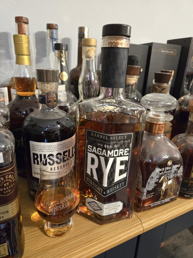 Close-up of a home whiskey bar with bottles of Sagamore Rye Barrel Select, Russell’s Reserve bourbon, and Rock Hill Farms on a wooden countertop, with a Glencairn tasting glass of amber whiskey in front.