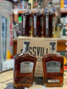 Two square-shouldered bottles of amber rye whiskey labeled “Rossville Union Master Crafted Straight Rye Whiskey” sit in the foreground on a wooden surface. Behind them is a cardboard box with more bottles packed inside, and additional upright bottles of the same whiskey blurred in the background on store shelves. The scene is inside a liquor store with other products faintly visible on shelves behind the display.