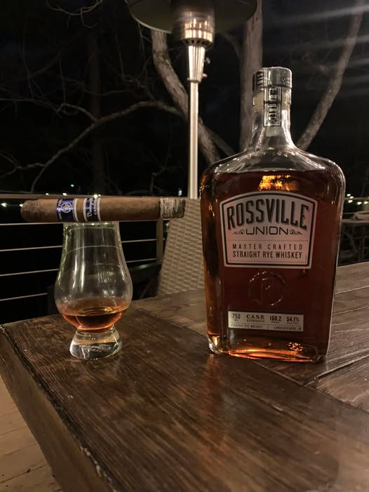 A nighttime outdoor scene showing a bottle of Rossville Union straight rye whiskey standing on a wooden table beside a short whiskey glass with amber liquid. A lit cigar rests across the rim of the glass. In the background are bare tree branches and a tall outdoor patio heater.