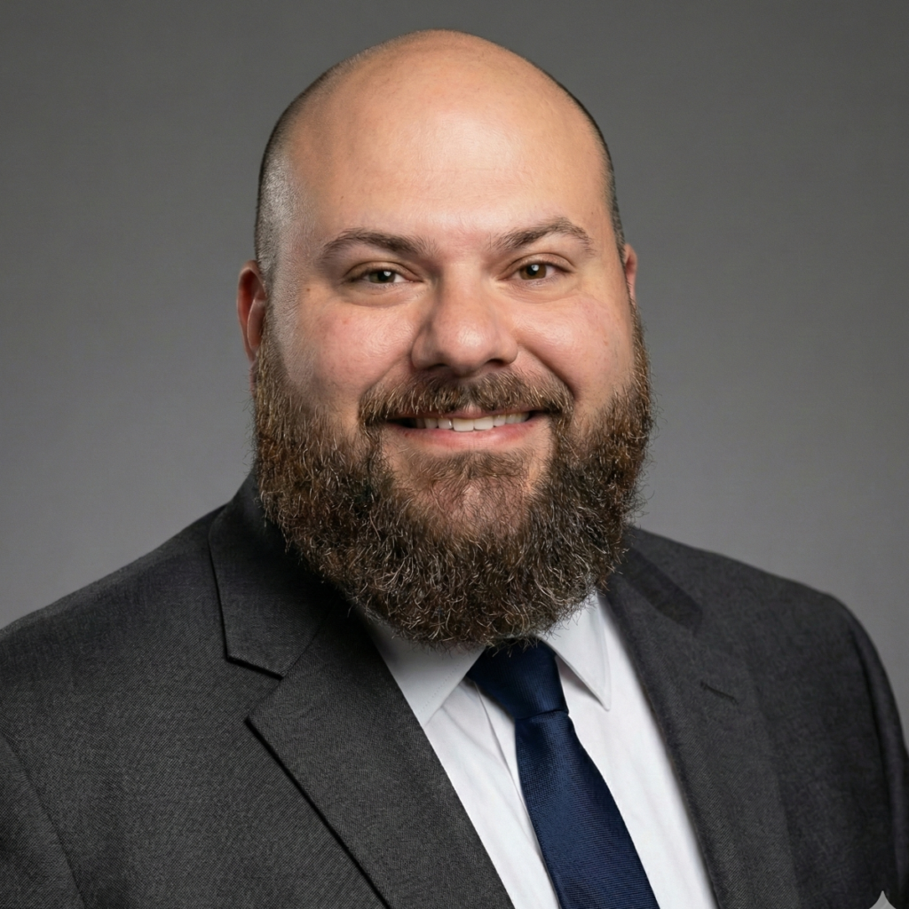 "Professional headshot of Drew Fowler smiling in a dark suit and tie with a groomed beard against a solid grey background.