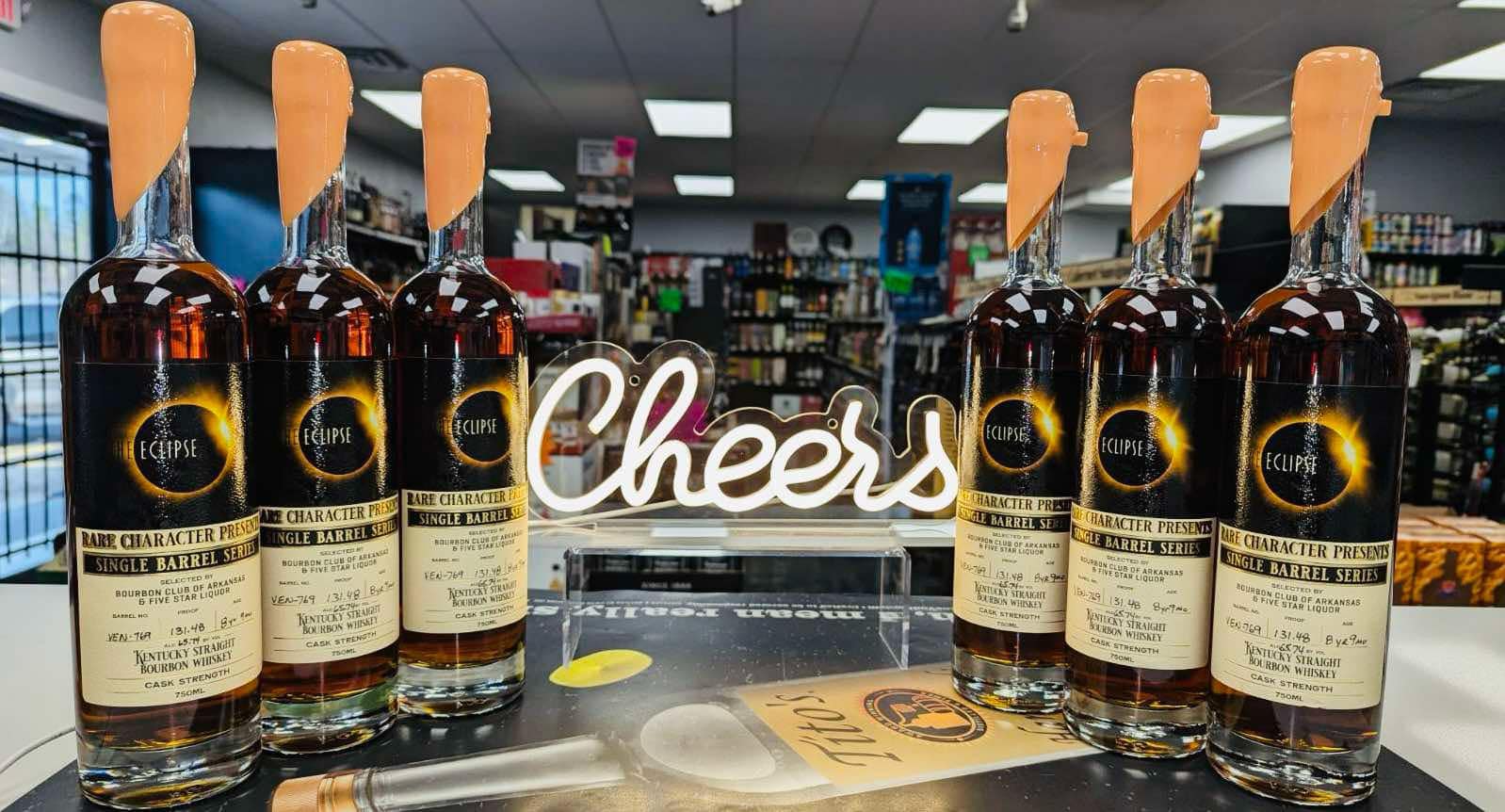 Six Eclipse Kentucky straight bourbon whiskey bottles from the Rare Character Presents Single Barrel Series lined up on a liquor store counter, with a lit ‘Cheers’ sign in the center and shelves of spirits in the background.