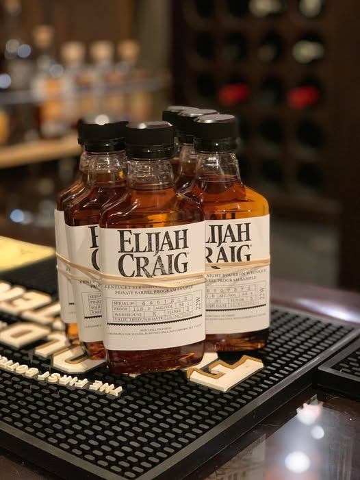 Close-up of several small Elijah Craig bourbon whiskey sample bottles with black caps, filled with amber liquid and wrapped in white “Elijah Craig” labels, grouped together on a black rubber bar mat atop a glossy bar counter, with a softly blurred background of liquor bottles and bar shelving suggesting a whiskey tasting or bourbon flight setup in a dimly lit bar or distillery tasting room.