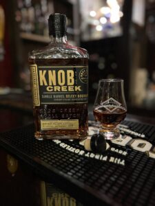 Close-up photo of a Knob Creek Single Barrel Select Kentucky Straight Bourbon Whiskey bottle on a black rubber bar mat, with a small Glencairn-style tasting glass filled with amber bourbon beside it. The bottle label reads “KNOB CREEK” and “Single Barrel Select Bourbon,” and the whiskey inside glows warm copper under bar lighting. The glass shows a bourbon club-style logo and highlights the rich color of the pour. In the foreground, two small round objects rest on the mat, while the background is softly blurred with shelves of liquor bottles and bokeh lights, creating a cozy cocktail bar atmosphere.