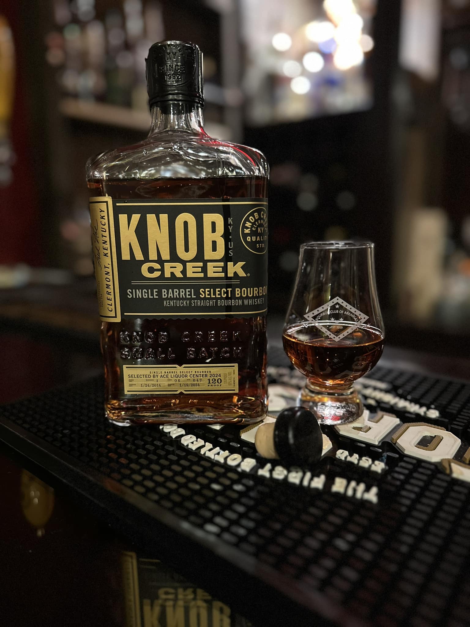 Close-up photo of a Knob Creek Single Barrel Select Kentucky Straight Bourbon Whiskey bottle on a black rubber bar mat, with a small Glencairn-style tasting glass filled with amber bourbon beside it. The bottle label reads “KNOB CREEK” and “Single Barrel Select Bourbon,” and the whiskey inside glows warm copper under bar lighting. The glass shows a bourbon club-style logo and highlights the rich color of the pour. In the foreground, two small round objects rest on the mat, while the background is softly blurred with shelves of liquor bottles and bokeh lights, creating a cozy cocktail bar atmosphere.