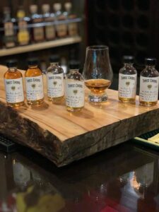 A wooden tasting board holding five small clear sample vials of Three Chord bourbon whiskey, each with a white label and handwritten notes, arranged in a row. In the center of the row behind the bottles is a filled whiskey tasting glass with amber liquid. The background shows blurred liquor store shelves with larger bottles on display.