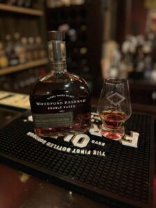 Woodford Reserve Double Oaked bourbon bottle (Barrel Finish Select, Ace Liquor/BCA Club Pick) next to a Glencairn glass with amber whiskey on a bar mat, with liquor shelves blurred in the background.