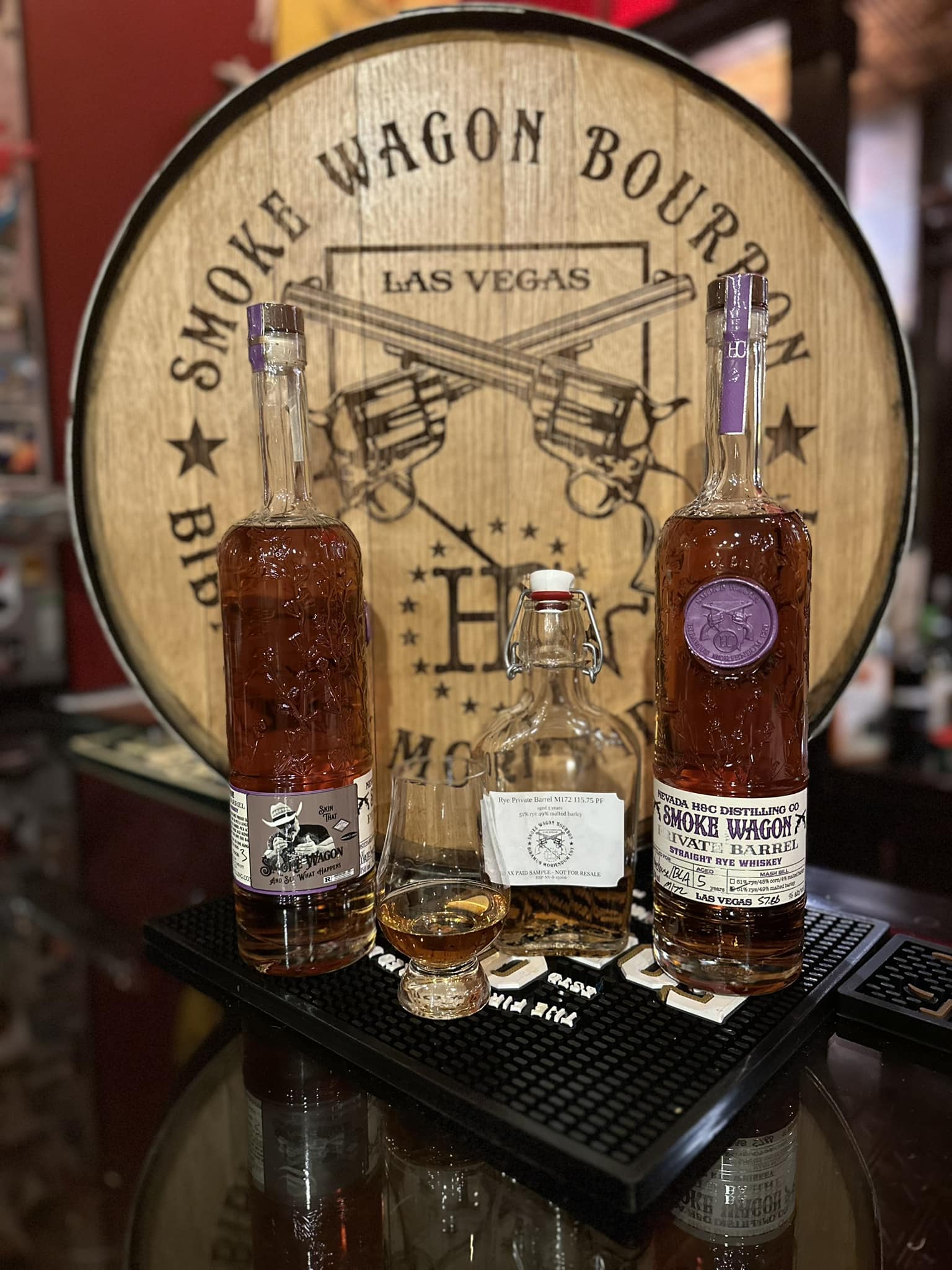 Three bottles of Smoke Wagon whiskey arranged on a black bar mat on a counter in front of a large wooden barrel top branded “Smoke Wagon Bourbon Las Vegas.” Two tall bottles of amber-colored Smoke Wagon bourbon stand on either side of a smaller clear sample flask with a swing-top cap. In front of the bottles is a filled whiskey tasting glass with amber liquid. The background shows part of the wooden barrel display and blurred store or tasting room elements.