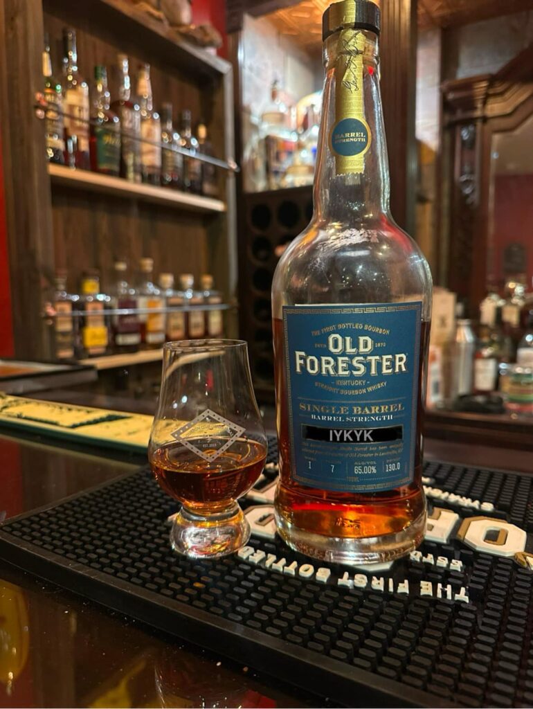 Old Forester Single Barrel Barrel Strength Kentucky straight bourbon whiskey bottle (blue label, “IYKYK,” 65.00% ABV / 130 proof) standing on a bar counter next to a Glencairn tasting glass filled with amber bourbon; warm, dimly lit whiskey bar background with shelves of liquor bottles softly blurred, highlighting a premium bourbon tasting and whiskey flight setting.