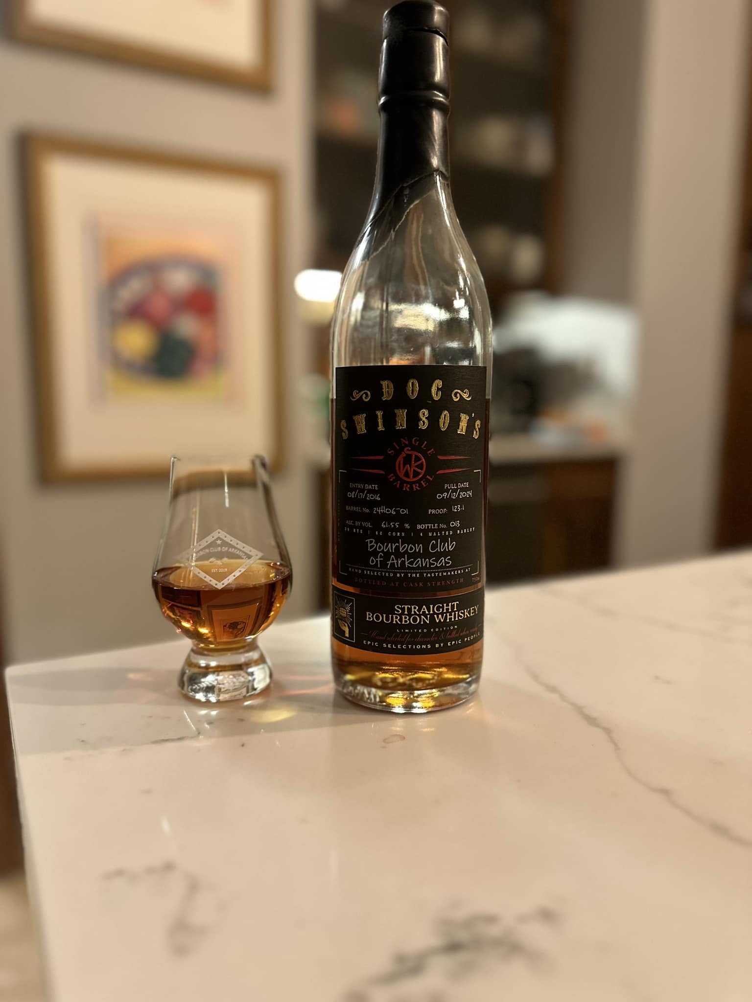 Doc Swinson’s Single Barrel Straight Bourbon Whiskey bottle standing on a white marble countertop beside a small Glencairn tasting glass filled with amber bourbon; close-up indoor bar scene with soft, blurred background artwork and shelving, highlighting the bourbon label and rich whiskey color for a bourbon tasting or whiskey review.