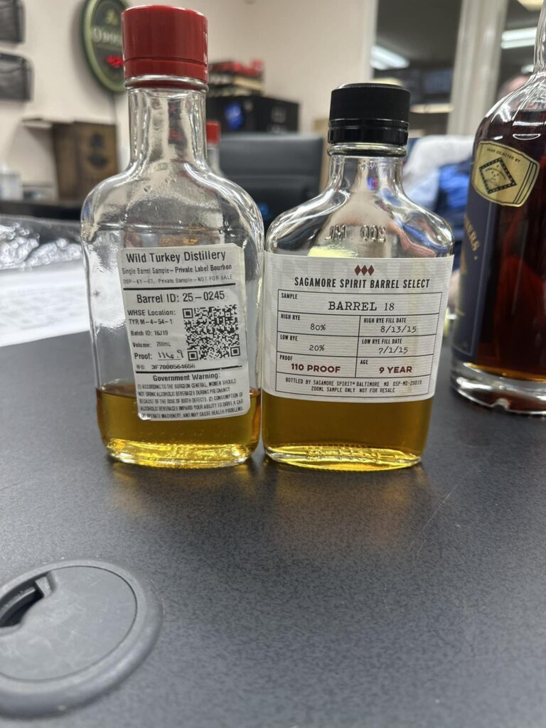 Close-up of two 200 ml whiskey sample bottles on a dark counter, partially filled with golden amber spirit—left is a Wild Turkey Distillery single barrel private label bourbon sample (red cap), right is a Sagamore Spirit Barrel Select high-rye sample labeled Barrel 18 (black cap).
