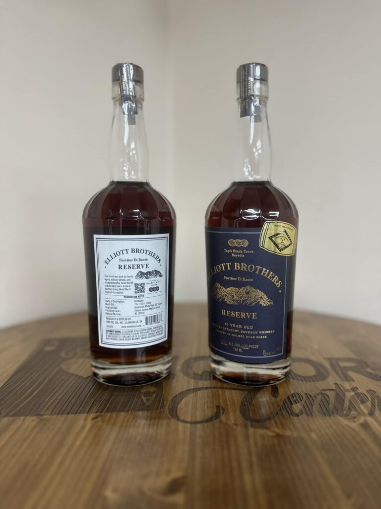 Two Elliott Brothers Reserve bourbon whiskey bottles standing side by side on a wooden tabletop, photographed indoors against a plain light-colored wall. The bottle on the left shows the back label—white with black text—reading ‘Elliott Brothers Reserve’ with production notes and a barcode visible through the clear glass; the bourbon inside is a deep amber-brown. The bottle on the right shows the front label—dark navy with gold lettering and a mountain illustration—reading ‘Elliott Brothers Reserve, 10 Year Old, Kentucky Straight Bourbon Whiskey,’ with a small gold ‘hand selection’ sticker near the top. Both bottles have silver caps and are centered in the frame, with the wood grain and faint branding on the table visible in the foreground.