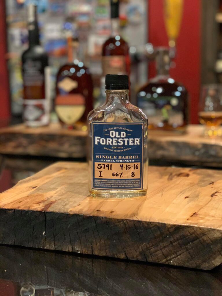 Old Forester Single Barrel Barrel Strength Kentucky Straight Bourbon Whiskey bottle with a black cap, photographed front-on on a thick live-edge wooden slab table. The blue and cream label reads ‘Old Forester’ and ‘Single Barrel Barrel Strength,’ with handwritten barrel details including ‘5791,’ date ‘4-15-16,’ and ‘66%’ (approx. 132 proof). In the softly blurred background is a home bar or whiskey shelf with several other bourbon bottles and a glass of amber whiskey, creating a warm, cozy tasting-room atmosphere.