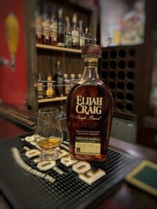 Elijah Craig Single Barrel Private Barrel Kentucky Straight Bourbon whiskey bottle on a bar counter beside a Glencairn tasting glass with a small pour of amber bourbon; liquor shelves with assorted bottles blur in the background, creating a warm, cozy bourbon bar and whiskey tasting atmosphere.