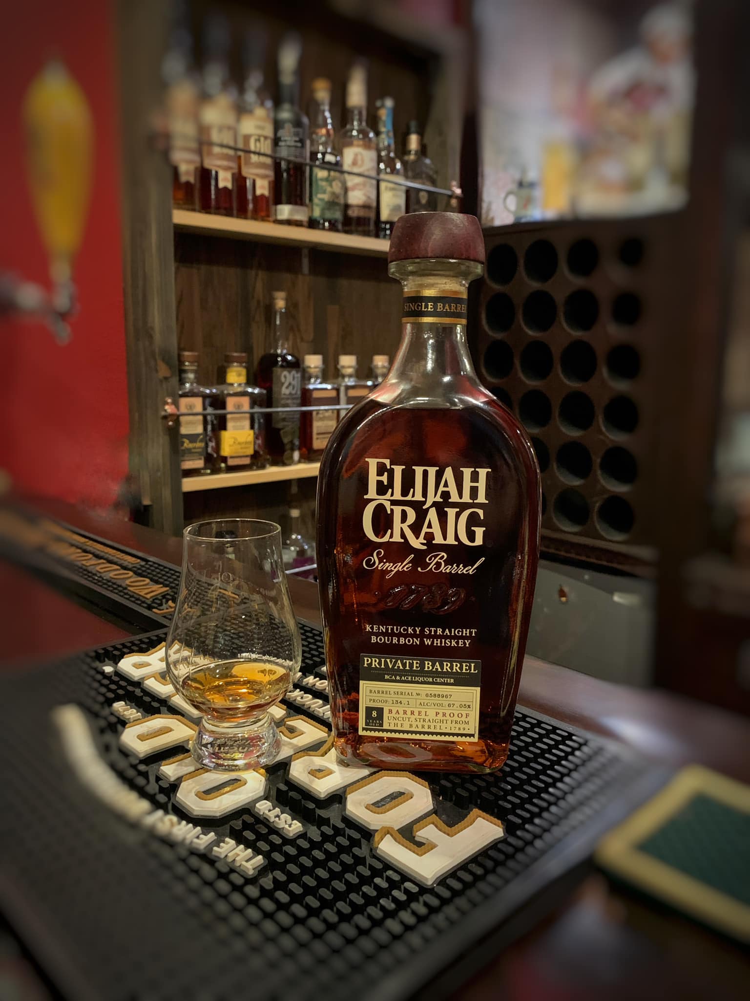 Elijah Craig Single Barrel Private Barrel Kentucky Straight Bourbon whiskey bottle on a bar counter beside a Glencairn tasting glass with a small pour of amber bourbon; liquor shelves with assorted bottles blur in the background, creating a warm, cozy bourbon bar and whiskey tasting atmosphere.