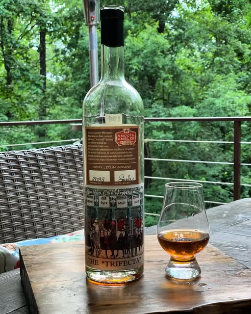 Smooth Ambler The Trifecta bourbon bottle beside a Glencairn-style whiskey glass with amber bourbon on a wooden patio table, trees in the background.