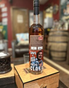 Close-up of a Smooth Ambler Spirits Old Scout bourbon bottle filled with amber whiskey, placed on a wooden box in a cozy bar or tasting room with blurred barrels in the background; the front label says ‘Save the neck pour for me, Clark’ and shows an illustrated man.