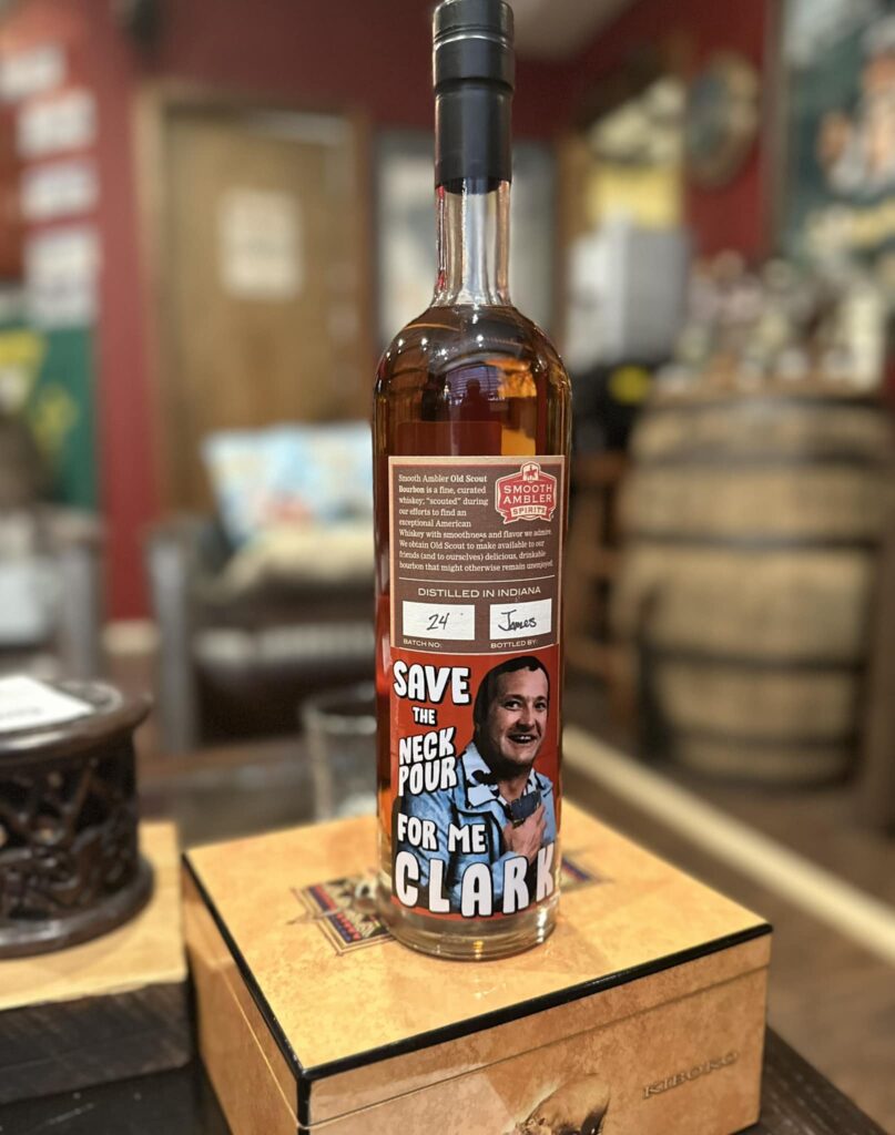 Close-up of a Smooth Ambler Spirits Old Scout bourbon bottle filled with amber whiskey, placed on a wooden box in a cozy bar or tasting room with blurred barrels in the background; the front label says ‘Save the neck pour for me, Clark’ and shows an illustrated man.