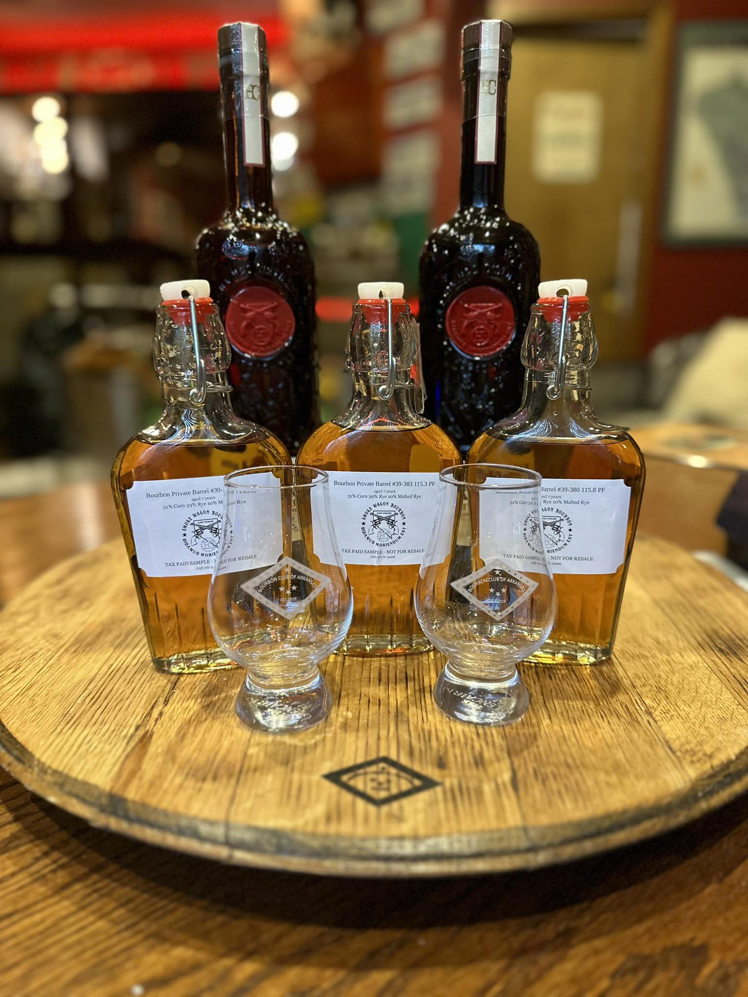 A portrait-style photo of two tall, slender bottles of Smoke Wagon bourbon whiskey with amber-colored liquid, standing side by side on a flat surface. Each bottle has a decorative label featuring intricate text and artwork. The background is blurred, showing what looks like a store or tasting room setting with warm lighting behind the bottles. The overall focus is on the rich amber color of the whiskey and the label details.