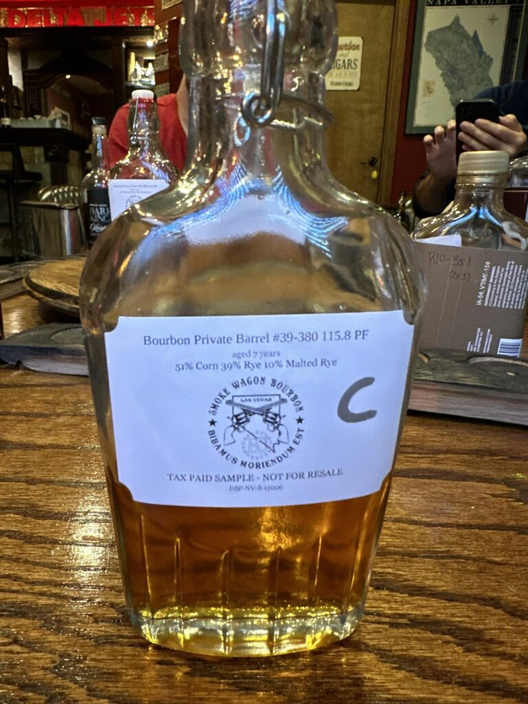 A clear glass sample bottle of Smoke Wagon Bourbon sitting on a wooden bar or table with amber-colored bourbon inside. The bottle has a white label that reads “Bourbon Private Barrel #39-380 115.8 PF, aged 7 years, 51% Corn 39% Rye 10% Malted Rye,” with the Smoke Wagon Bourbon logo and text “Tax Paid Sample — Not For Resale.” Behind the bottle are other bottles and a bar-like setting with people partially visible in the background.