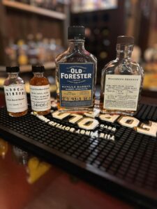 Close-up photo of a bourbon tasting setup on a bar. Two small sample bottles labeled ‘Doc Stetson’s Single Barrel Sample’ and a ‘Straight Bourbon Whiskey’ sample sit beside two full-size Kentucky bourbon bottles: Old Forester Single Barrel Barrel Strength and Woodford Reserve Double Oaked Personal Selection. The bottles are lined up on a black rubber bar mat with raised lettering, with a softly blurred back-bar of liquor bottles in the background, suggesting a whiskey flight or single-barrel selection tasting.