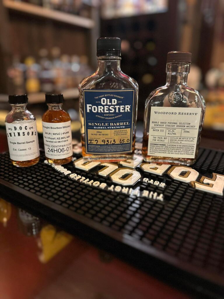 Close-up photo of a bourbon tasting setup on a bar. Two small sample bottles labeled ‘Doc Stetson’s Single Barrel Sample’ and a ‘Straight Bourbon Whiskey’ sample sit beside two full-size Kentucky bourbon bottles: Old Forester Single Barrel Barrel Strength and Woodford Reserve Double Oaked Personal Selection. The bottles are lined up on a black rubber bar mat with raised lettering, with a softly blurred back-bar of liquor bottles in the background, suggesting a whiskey flight or single-barrel selection tasting.
