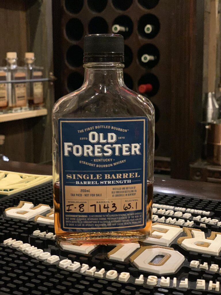 A small clear sample bottle of Old Forester bourbon sitting on a flat surface inside a liquor store. The bottle contains amber-colored whiskey and carries a label identifying it as an Old Forester expression (a classic Kentucky straight bourbon brand). Behind it are blurred shelves of other spirits and store lighting, giving context to a tasting or retail setting.