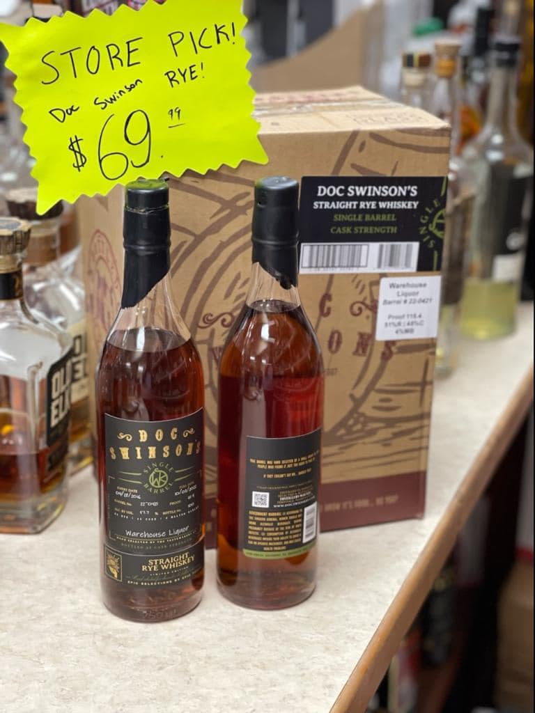Two bottles of amber-colored rye whiskey standing upright on a store counter in front of a cardboard box. Both bottles have dark labels reading “Doc Swinson’s Straight Rye Whiskey.” Behind them is a bright yellow starburst sign that says “STORE PICK! Doc Swinson RYE! $69.99.” Shelves of other liquor bottles are visible in the blurred background.