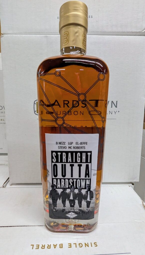 Close-up product photo of a Bardstown Bourbon Company whiskey bottle with amber bourbon and a beige cap, featuring a black-and-white label that reads ‘Straight Outta Bardstown,’ photographed in front of stacked Bardstown Bourbon Company boxes.