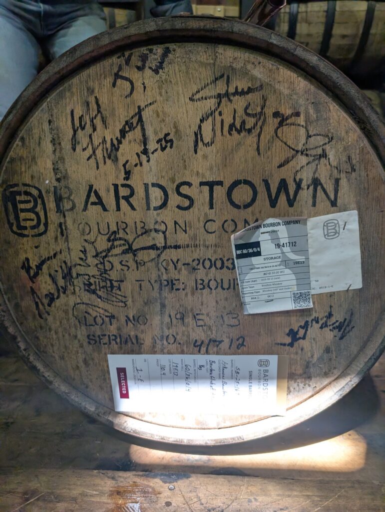 Close-up of a Bardstown Bourbon Company oak whiskey barrel head with handwritten rickhouse markings and inventory stickers, photographed in a bourbon barrel warehouse.