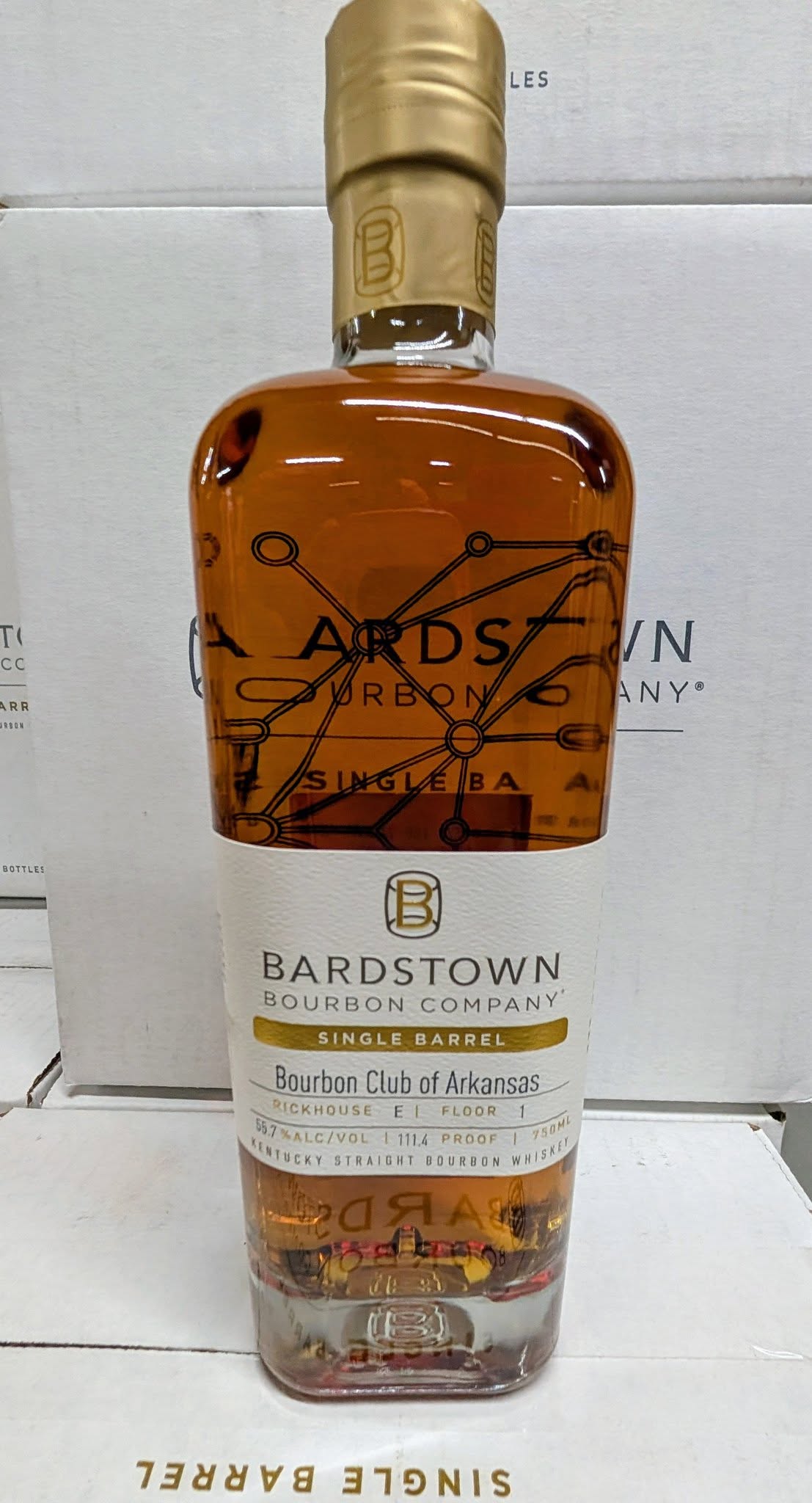 Front view of a Bardstown Bourbon Company Single Barrel Kentucky straight bourbon whiskey bottle with amber liquid, gold foil neck, and label reading ‘Bourbon Club of Arkansas,’ ‘Rickhouse E1, Floor 1,’ 55.7% ABV, 111.4 proof, 750 ml, photographed against stacked white Bardstown Bourbon Company boxes.