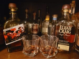 Close-up of a home bar shelf with several whiskey and bourbon bottles and two empty glass tumblers in the foreground. The right bottle label reads ‘Elijah Craig Single Barrel Kentucky Straight Bourbon Whiskey Private Barrel,’ and the left bottle shows an illustrated label with the text ‘Liquorem Veritas.’ Amber-colored spirits are visible in the bottles, and warm indoor lighting highlights the glass and wooden caps, suggesting a bourbon tasting or whiskey bar setup.