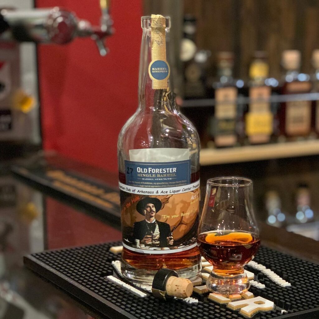 Old Forester Single Barrel Barrel Strength bourbon bottle on a bar mat beside a Glencairn glass filled with amber whiskey and a curled orange peel; the bottle has a blue Old Forester label and a custom illustrated sticker, with the cork lying in front, while liquor bottles and bar equipment blur in the warm, dim background of a whiskey bar.