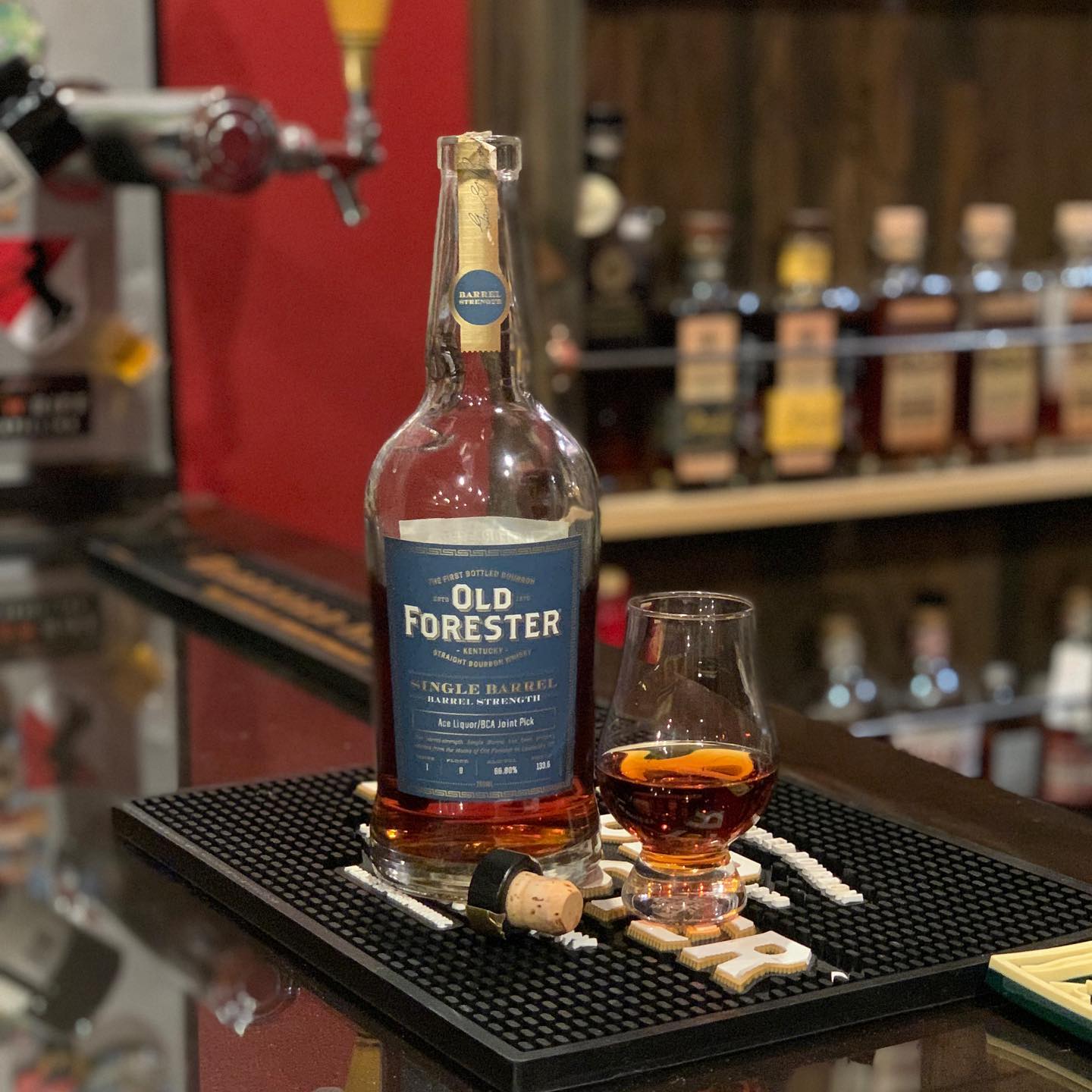 Old Forester Single Barrel Barrel Strength Kentucky straight bourbon bottle on a bar counter beside a Glencairn tasting glass filled with amber whiskey; cork and bottle stopper resting on a black rubber bar mat, with a softly blurred background of liquor bottles on shelves and a beer tap, capturing a premium bourbon tasting and bar setup.