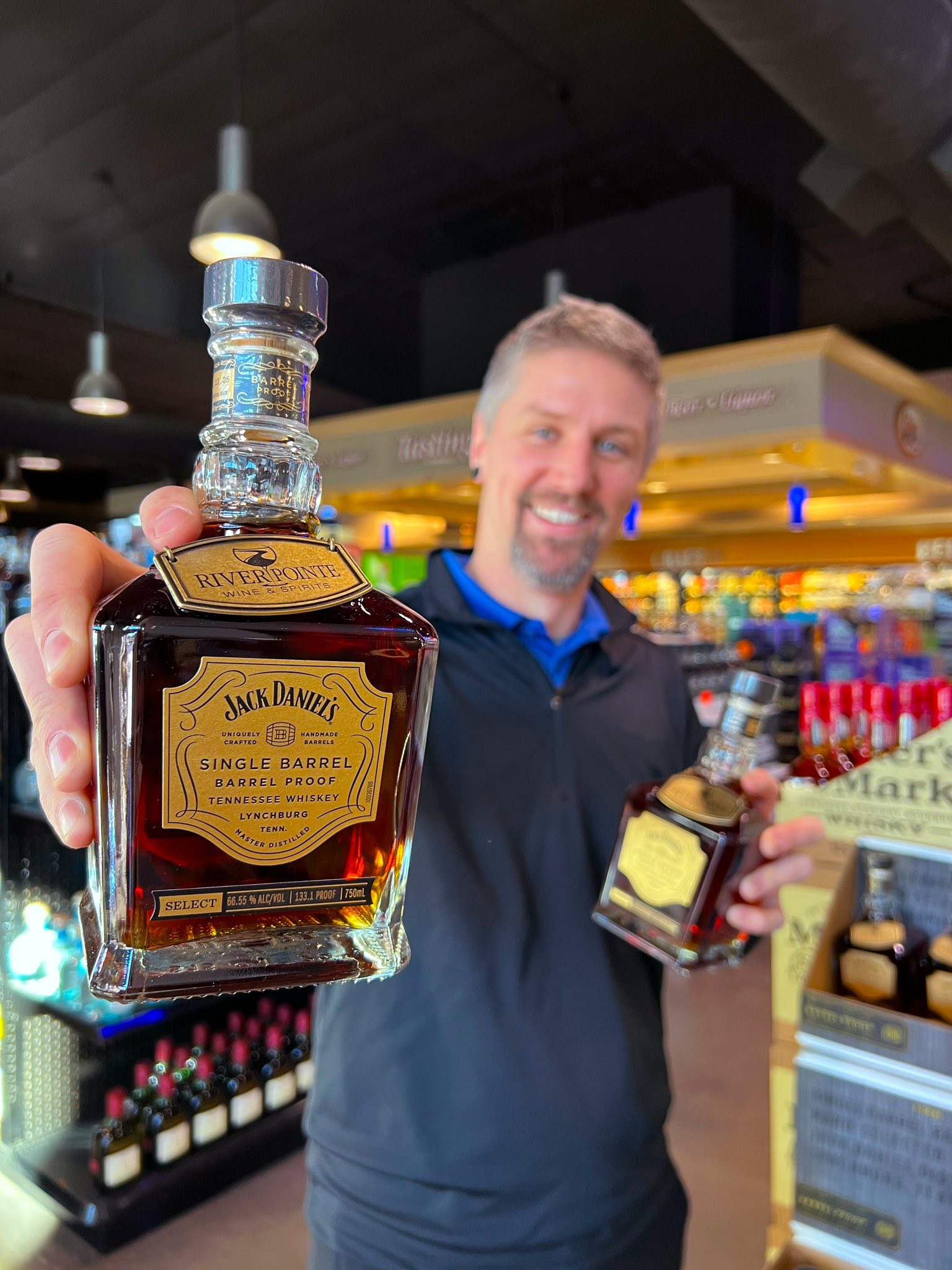 Man holding a Jack Daniel’s Single Barrel Barrel Proof Tennessee Whiskey bottle in a liquor store aisle, with shelves of spirits and wine in the background.