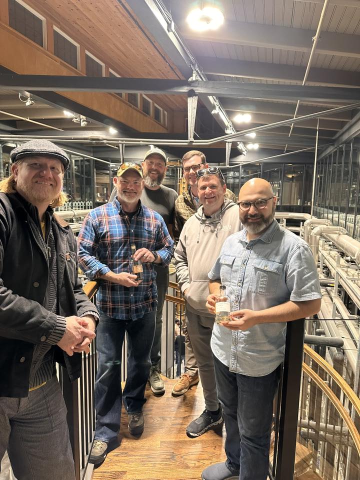 Group of friends on a distillery tour inside a whiskey distillery, holding tasting glasses on an industrial catwalk above stainless steel production equipment.