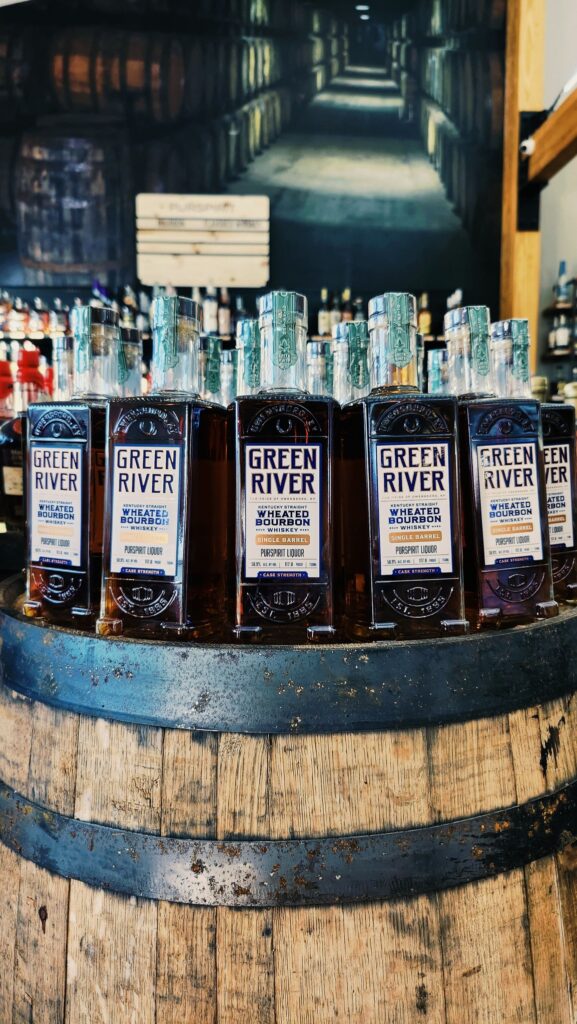 Green River Wheated Bourbon whiskey bottles displayed on a wooden barrel Purspirit Liquor, with barrel warehouse mural in background.